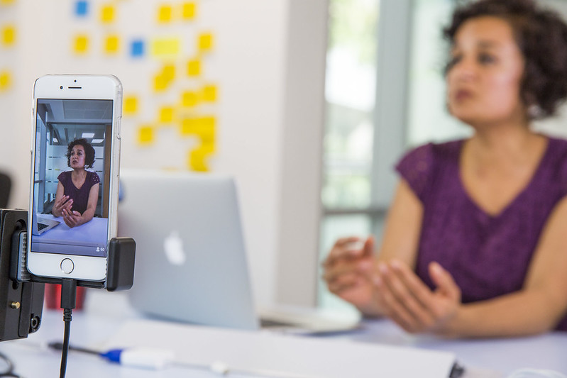 Woman sitting at a desk talking being filmed on a mobile phone.